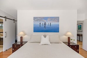 Ein Schlafzimmer mit einem großen Bett, zwei Nachttischen mit Lampen, einem Bild an der Wand und Blick zur Küche.