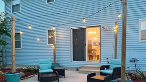 A blue house with a patio area featuring two wicker chairs with cushions, a small table, and string lights.