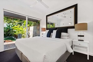 Ein modernes Schlafzimmer mit einem großen Bett, einem Deckenventilator, einem Nachttisch mit Lampe und Blick auf einen Garten durch eine Schiebetür.