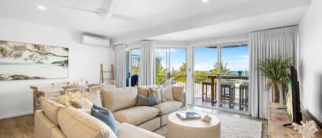 Un salon spacieux avec un grand canapé, une table basse et un téléviseur. Il y a un balcon avec vue sur l’océan.