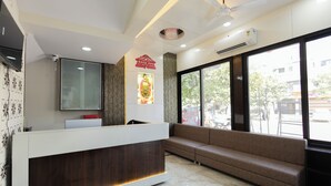 A reception area with a modern desk, a sofa, a television, and a large window offering a view of the street. - Hotel Basil Inn (Ujjain)