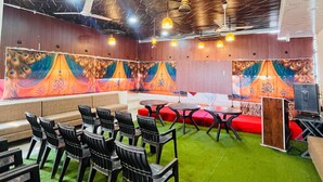 A room with a green carpet, black chairs, and a red sofa. There are colorful wall tapestries and a ceiling fan. - Hotel Basil Inn (Ujjain)