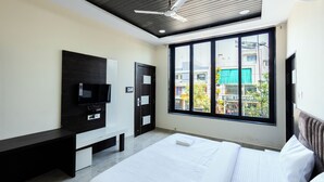 A modern bedroom with a large bed, a TV mounted on the wall, a ceiling fan, and a window with a city view. - Hotel Basil Inn (Ujjain)