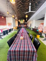 A long dining table with a plaid tablecloth, surrounded by black chairs, in a room with a high ceiling and hanging lights.