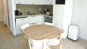 Een compacte keuken met witte kastjes, een magnetron, een spoelbak en een kleine eettafel met stoelen.