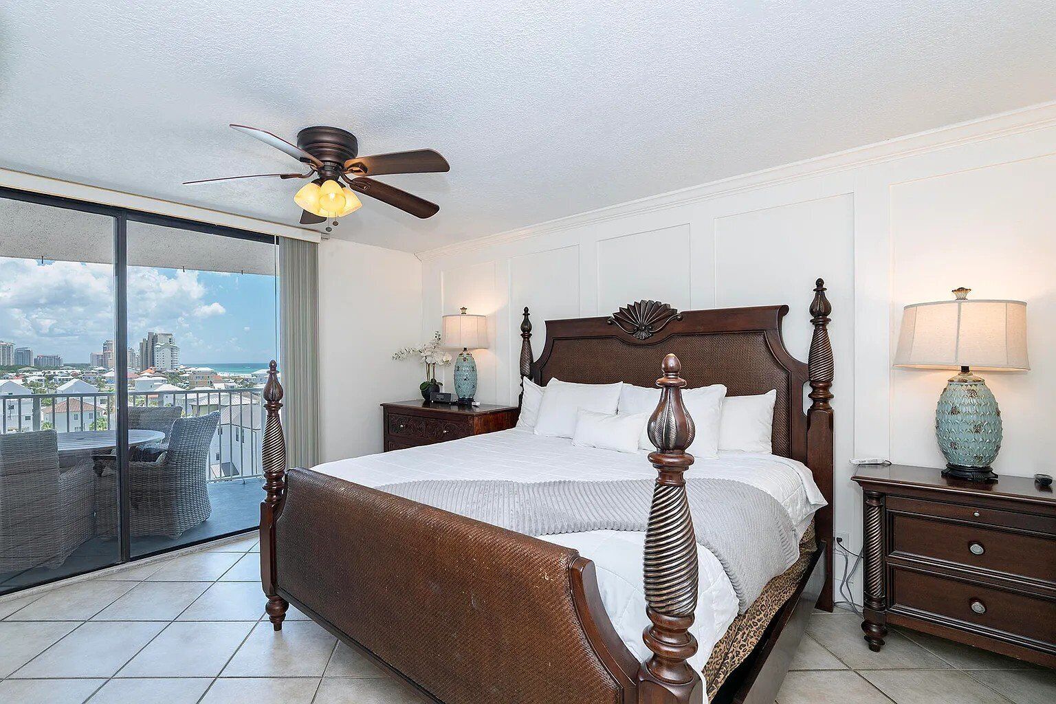 A spacious bedroom with a large bed, a ceiling fan, and a view of the city through a sliding glass door.