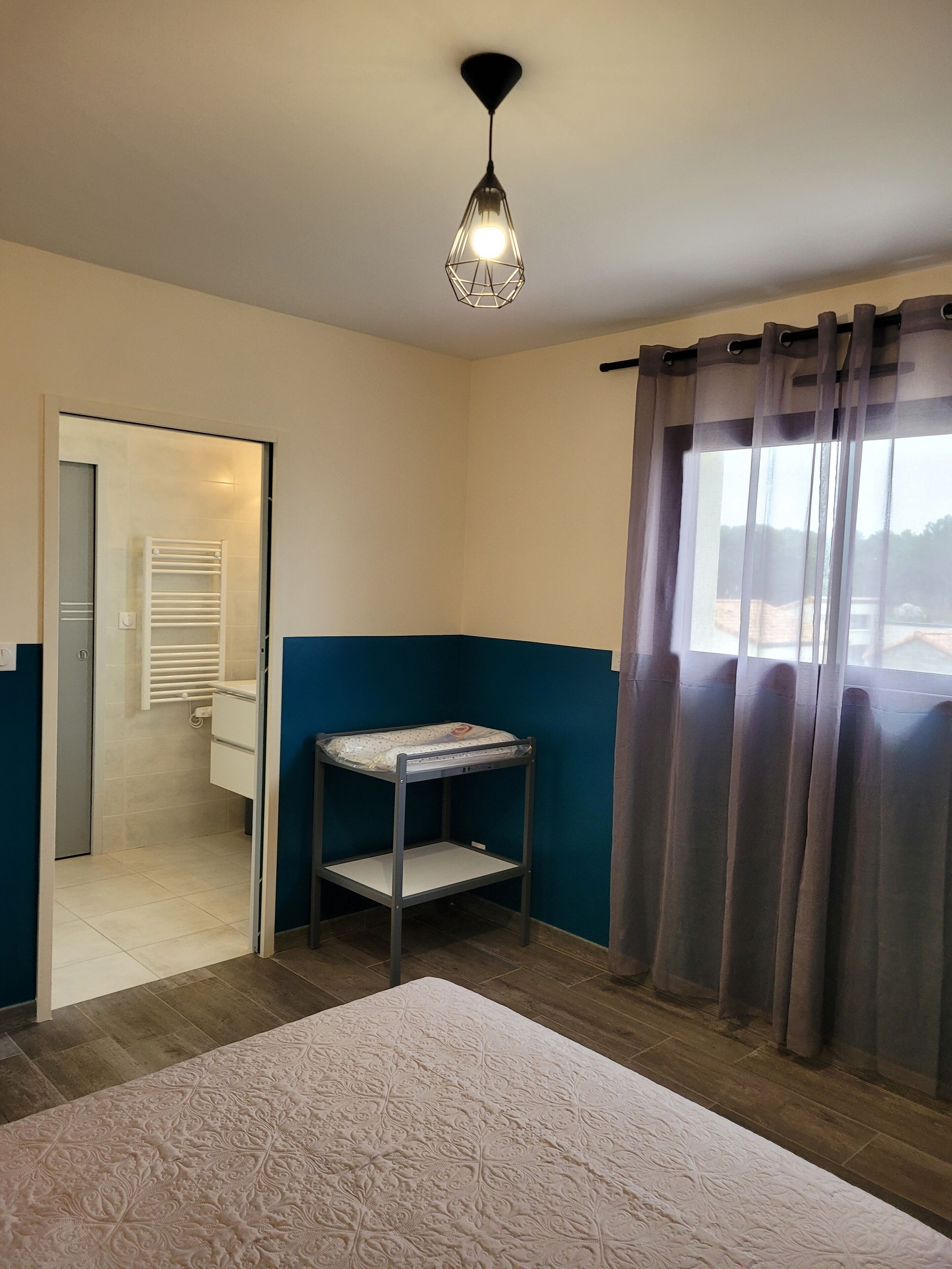 Une chambre à coucher avec un lit, un luminaire suspendu, une fenêtre avec des rideaux et une vue sur un bâtiment à l’extérieur.