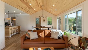 Un soggiorno moderno con soffitto in legno, un divano con cuscini, un'area pranzo con tavolo e sedie, e ampie finestre che offrono una vista.