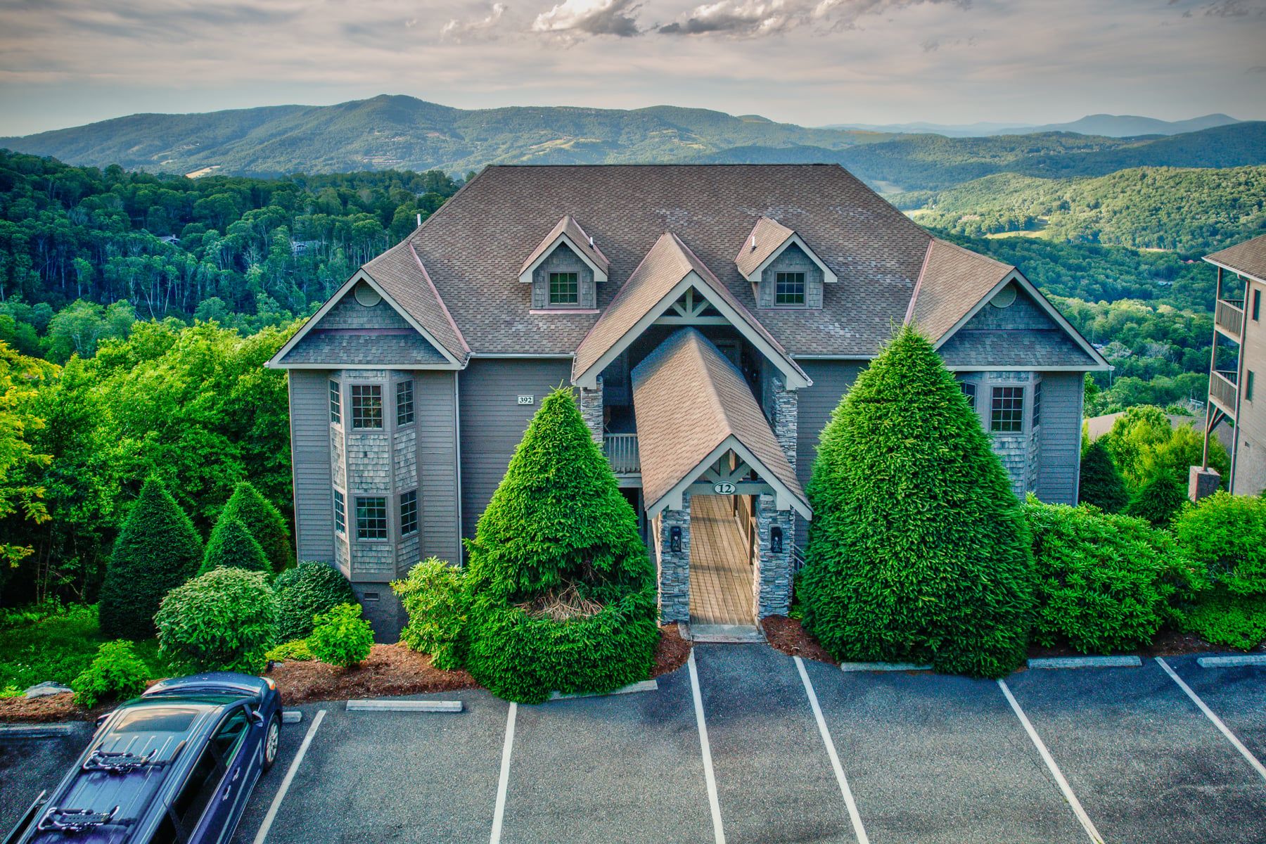 A two-story house with a steeply pitched roof, a front porch, and a driveway leading to a parking area.
