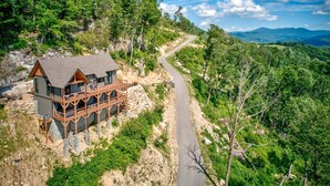 A cabin with a deck, surrounded by forested hills and a winding road. - Clouds in My Coffee at Eagles Nest- walk to Village amenities, view, hot tub, golden tee arcade (Banner Elk)