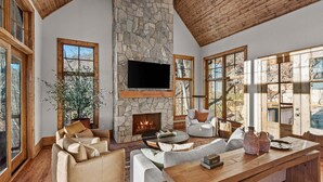 A spacious living room with a stone fireplace, comfortable seating, large windows, and a wooden ceiling. - Clouds in My Coffee at Eagles Nest- walk to Village amenities, view, hot tub, golden tee arcade (Banner Elk)