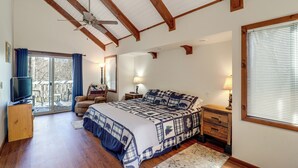 Quarto com uma cama grande, vigas de madeira, ventilador de teto, televisão, uma cadeira, mesa de cabeceira e janela com persianas.