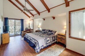 Een slaapkamer met een groot bed, houten balken, een plafondventilator, een televisie, een stoel, een nachtkastje en een raam met jaloezieën.