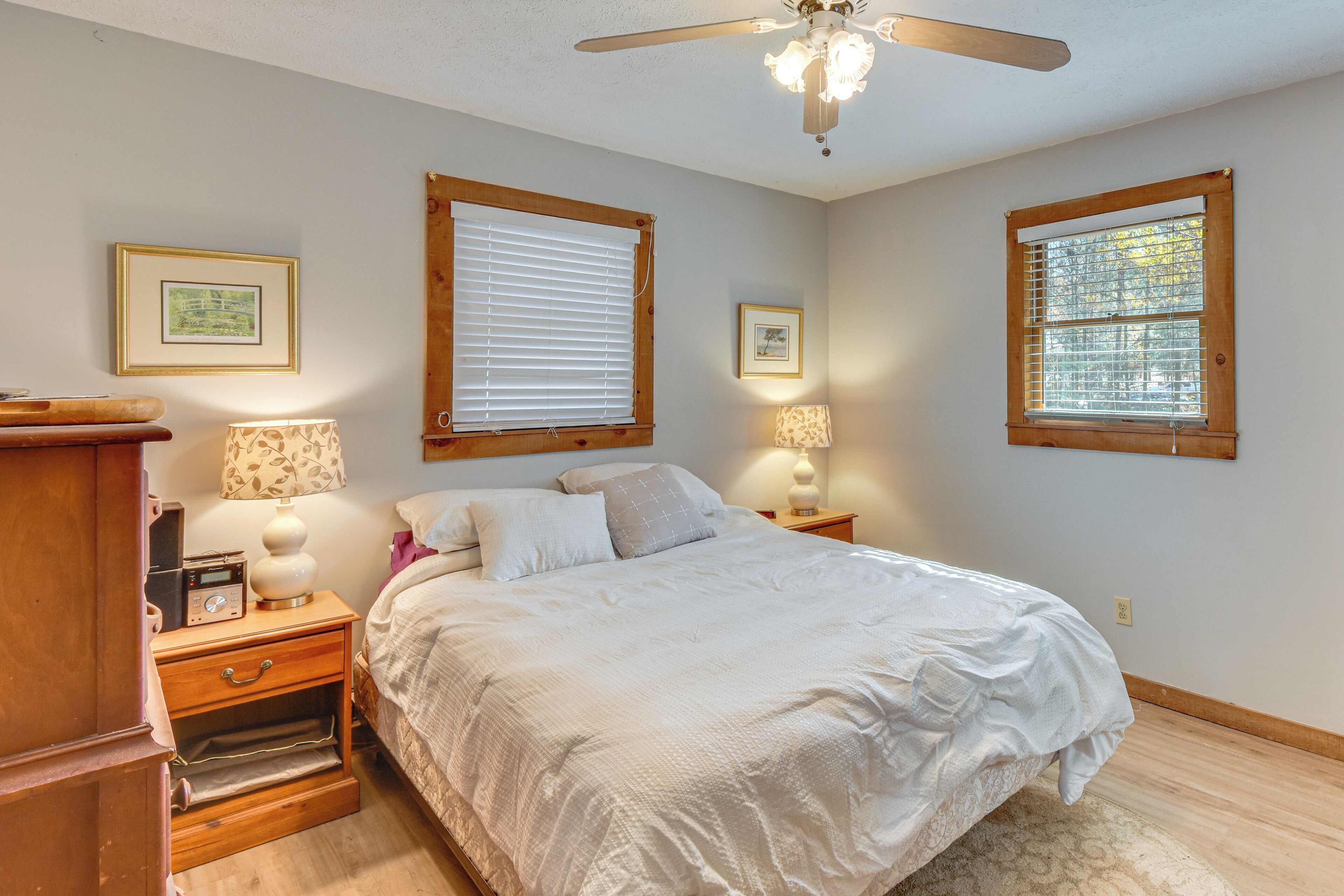 A bedroom with a bed, two bedside tables, a nightstand, a ceiling fan, and two windows with blinds.
