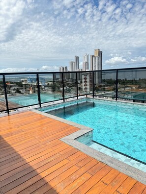 A rooftop pool with a wooden deck and a glass barrier, overlooking a cityscape with high-rise buildings.