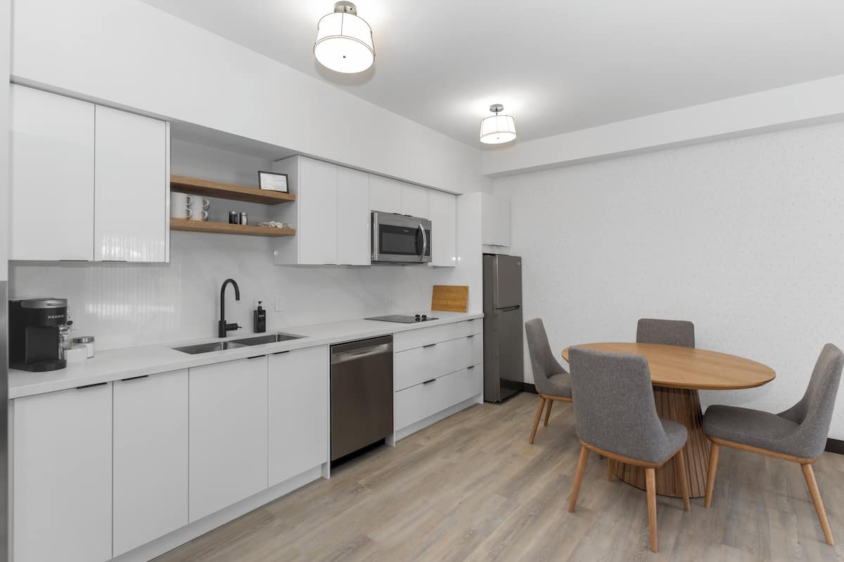 A modern kitchen with white cabinets, a stainless steel dishwasher, a microwave, and a refrigerator. There is a wooden dining table with four chairs.