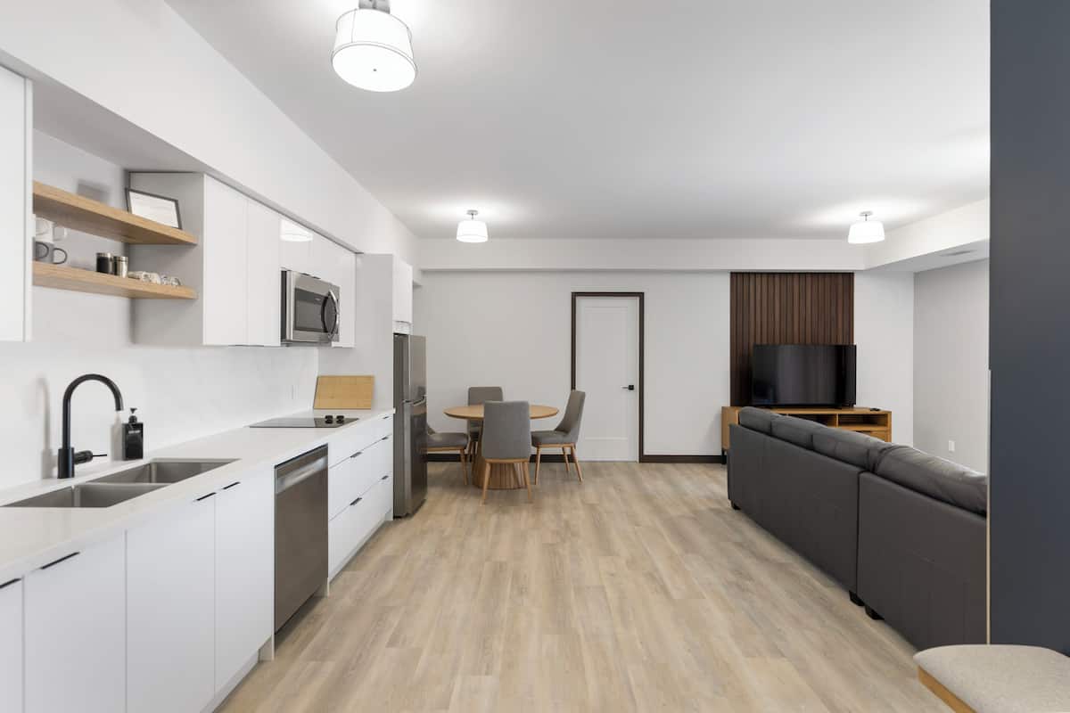 A modern kitchen with white cabinets, a microwave, and a dishwasher. There is a dining area with a table and chairs. A gray sofa and a TV are visible in the living area.
