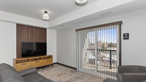 A modern living room with a flat-screen TV, wooden entertainment unit, grey sofas, and a view through a sliding glass door. - Lux Suites Extended Stay (Thunder Bay)