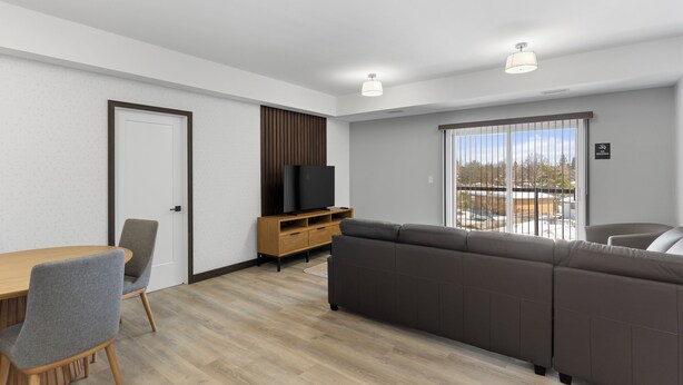 A modern living room with a wooden dining table, grey chairs, a brown sofa, a TV on a wooden stand, and a large window with a view. - Lux Suites Extended Stay (Thunder Bay)