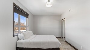 A bedroom with a bed, a window, a door, and a ceiling light fixture. - Lux Suites Extended Stay (Thunder Bay)