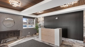 A modern reception area with a wooden ceiling, a reception desk, a dark wall with a circular mirror, and a window with a view of the outside. - Lux Suites Extended Stay (Thunder Bay)