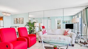 A modern living room with a white sofa, red armchairs, a glass coffee table, and a large mirror reflecting the room. - Relaxing Oceanfront Condo w/ Balcony, Pool & Direct Beach Access (New Smyrna Beach)