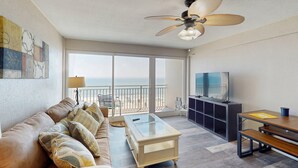 A spacious living area with a sofa, coffee table, and TV, overlooking a balcony with a view of the beach. - Private Balcony, Oceanfront Pool, Walk to Boardwalk & Pier (Daytona Beach)