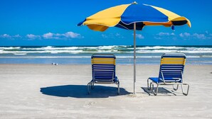 Two beach chairs under a blue and yellow umbrella on a sandy beach with the ocean in the background. - Private Balcony, Oceanfront Pool, Walk to Boardwalk & Pier (Daytona Beach)