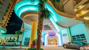 Ein moderner Hotel Eingang mit einer gewölbten Fassade, Palmen und einem Parkplatz mit Autos.