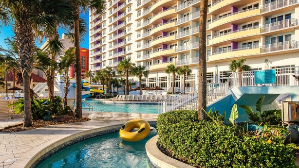 Ein Hotelpoolbereich mit einem runden Pool, Palmen und einem mehrstöckigen Gebäude mit Balkonen.