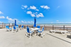 Un patio frente al mar con sillas y mesas blancas, sombrillas azules y vista al océano.