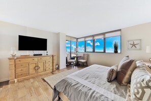 Un dormitorio con cama, televisor, cómoda y ventana con vista a palmeras.