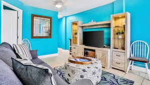A living room with a teal wall, a ceiling fan, a flat-screen TV, a wooden cabinet, and a sofa with patterned cushions. - Ground Floor Oceanfront Condo with Pools & Easy Beach Access (Daytona Beach)
