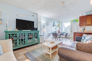 Ein Wohnzimmer mit Fernseher, eine Essecke mit Stühlen, ein Küchenbereich mit Schränken und Blick ins Freie.