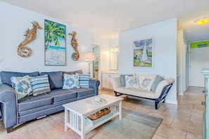 Ein Wohnzimmer mit einem blauen Sofa, einem weißen Couchtisch und zwei weißen Sesseln. An den Wänden befinden sich Kunstwerke, darunter ein Seepferdchen und ein Segelboot.
