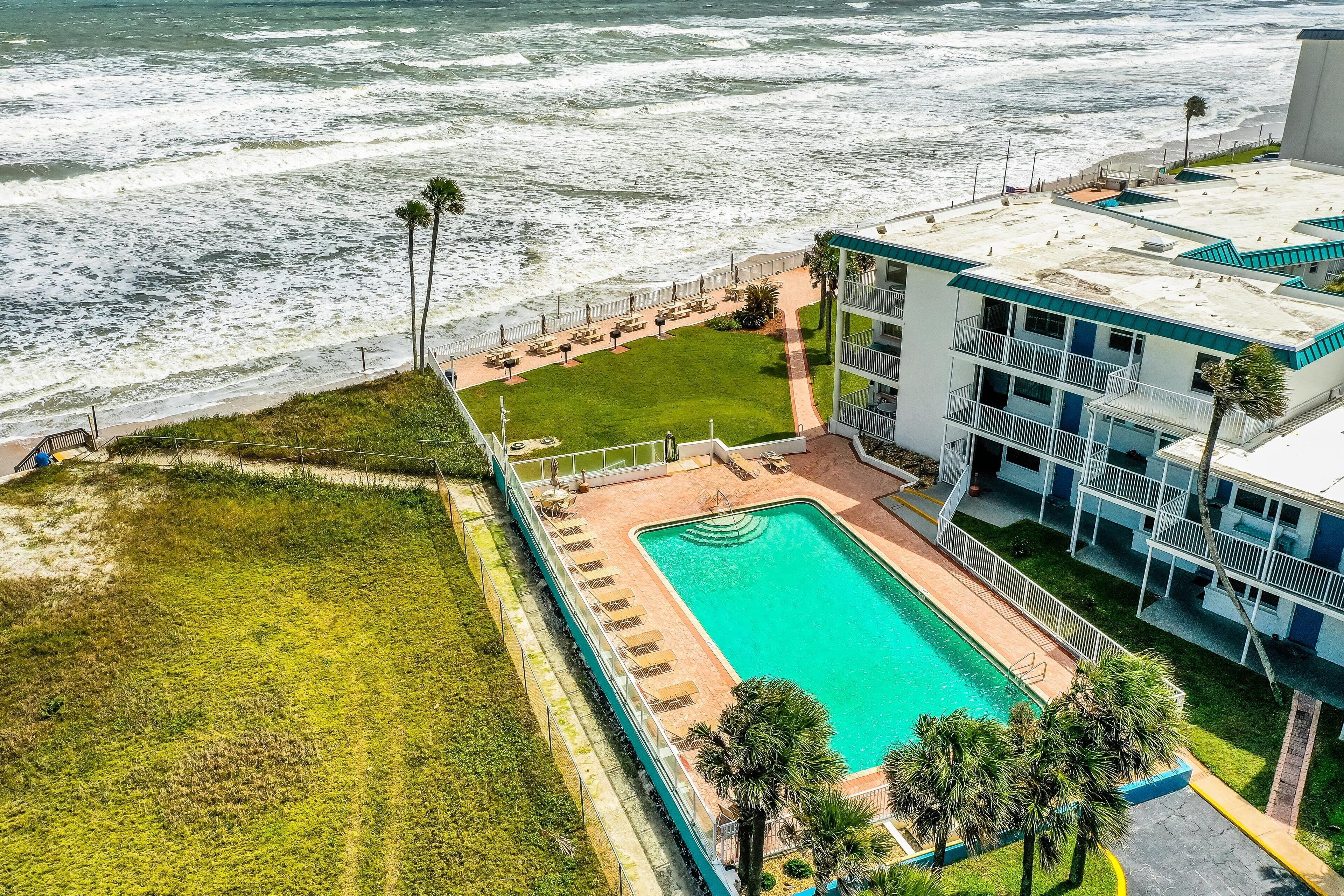 An aerial view of a hotel with a swimming pool, a grassy area, and a beachside view.