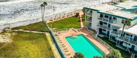 An aerial view of a hotel with a swimming pool, a grassy area, and a beachside view.