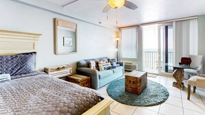 Habitación de hotel con cama, sofá, mesa de centro, sillón, ventilador de techo y ventana con cortinas.