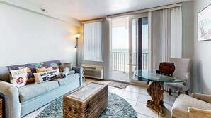Una sala de estar con un sofá, una mesa baja, una mesa con cubierta de vidrio y una silla. Hay una puerta corrediza de vidrio que da a un balcón.