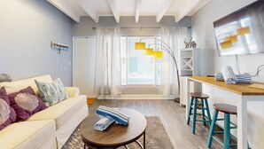 Ein modernes Wohnzimmer mit einer Couch, einem runden Couchtisch, einem Holztisch mit Hocker und einem großen Fenster mit Vorhängen.