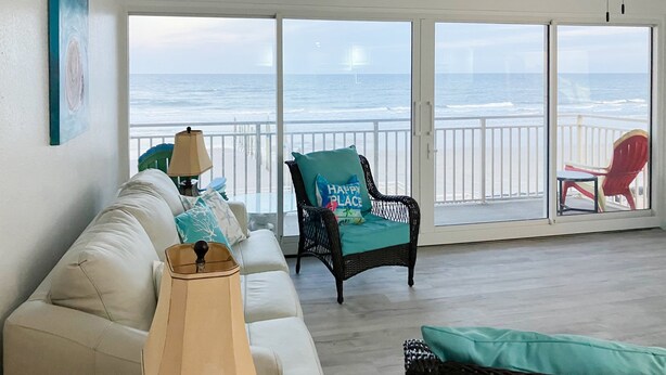 Ein Wohnzimmer mit einem weißen Sofa, einem Korbstuhl mit einem 'Happy Place'-Kissen, einer Lampe und Blick auf den Ozean durch große Schiebeglastüren.