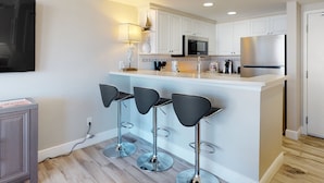 A modern kitchen with white cabinets, a stainless steel refrigerator, a microwave, and three black bar stools. - Oceanfront 12th-Floor Suite with Views, Pool & Prime Location (Daytona Beach)