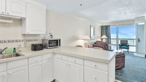 Eine moderne Küche mit weißen Schränken, einer Mikrowelle und einem Toaster. Ein Essbereich mit einer Couch und einem Sessel sowie ein Balkon mit Blick.