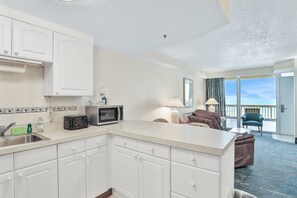 Eine moderne Küche mit weißen Schränken, einer Mikrowelle und einem Toaster. Ein Essbereich mit einer Couch und einem Sessel sowie ein Balkon mit Blick.