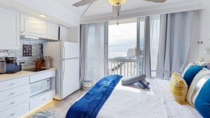 A hotel room with a balcony, a bed with blue and yellow pillows, a white ceiling fan, and a kitchen area with a microwave. - Chic Oceanfront Studio with Balcony, Pool & Direct Beach Access (Daytona Beach)