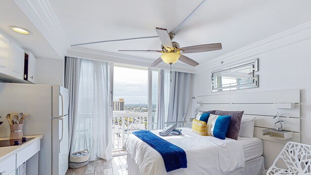 A hotel room with a bed, a desk, a chair, a ceiling fan, and a view of the city. - Chic Oceanfront Studio with Balcony, Pool & Direct Beach Access (Daytona Beach)