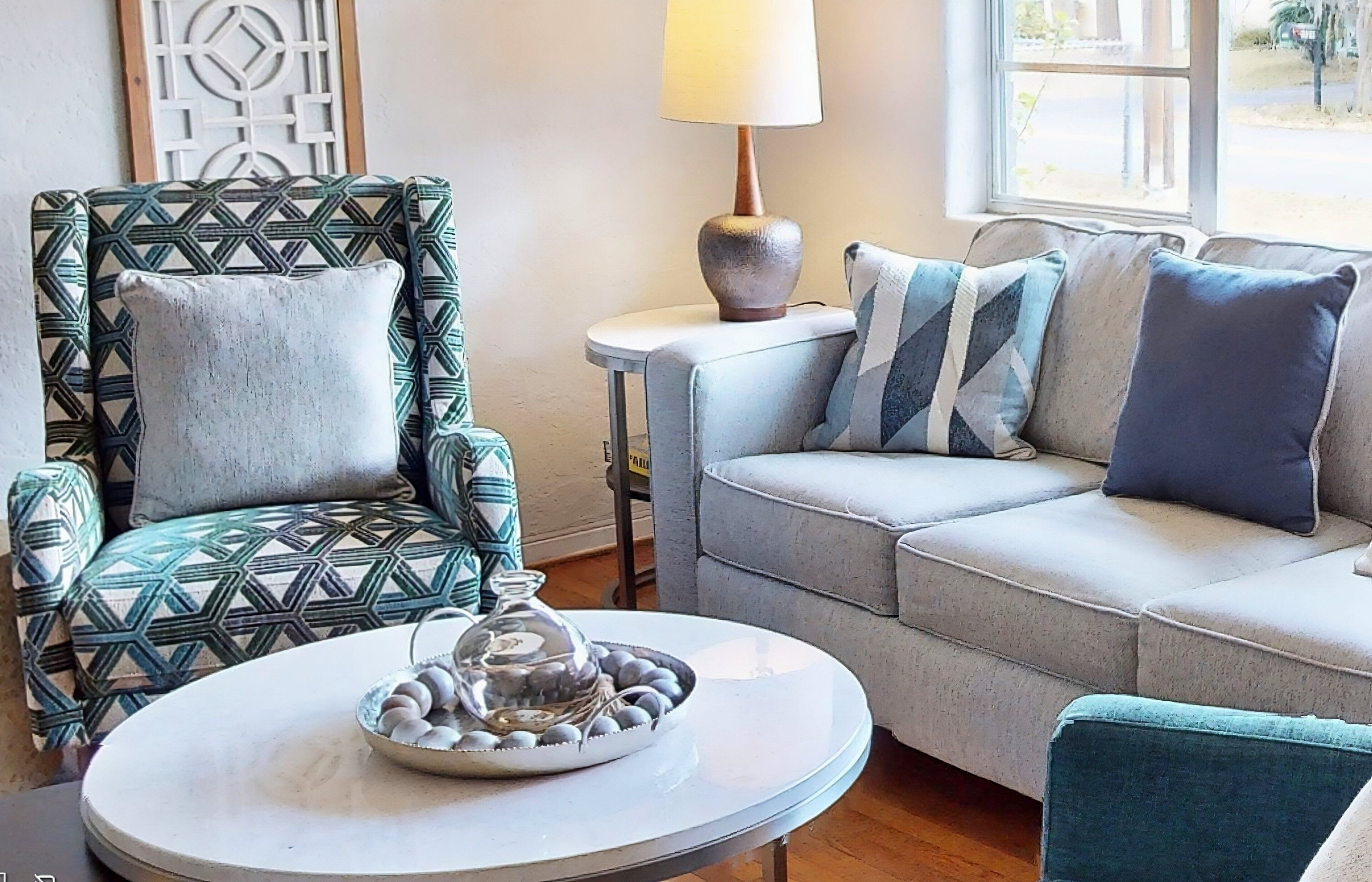 A living room with a patterned armchair, a sofa with cushions, a small table with a lamp, and a decorative glass bowl on the table.