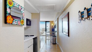 A narrow hallway with a kitchen area on the left, featuring white cabinets and a microwave. A trash bin is visible on the floor. The hallway leads to a dining area with a table and chairs.