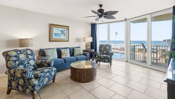 Ein Wohnzimmer mit einem blauen Sofa, gemusterten Sesseln, einem Couchtisch und einem Deckenventilator. Ein großes Fenster bietet Blick auf den Strand.