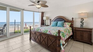 Ein Schlafzimmer mit Bett, Nachttisch, Lampe und Deckenventilator. Ein großes Fenster bietet Blick auf den Strand.